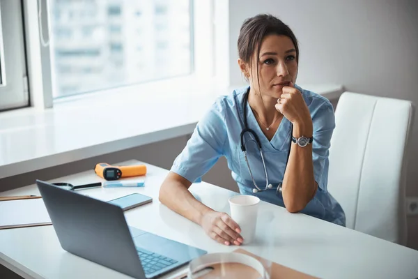 Encantadora doctora bebiendo café y usando computadora portátil en la clínica —  Fotos de Stock