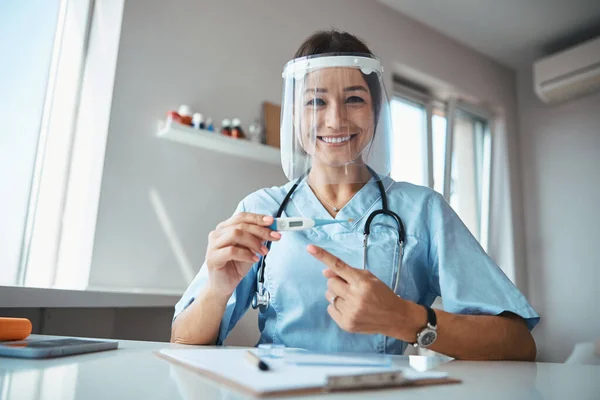 디지털 온도계를 가리키는 마스크를 쓰고 있는 쾌활 한 의사 — 스톡 사진