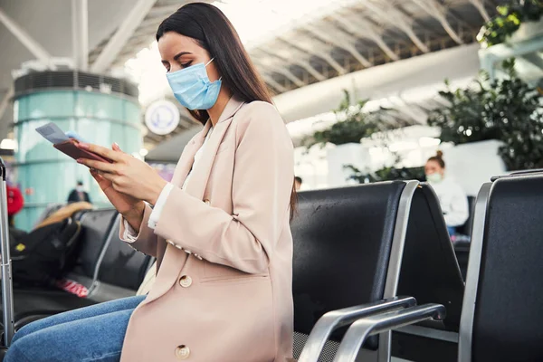 Stílusos nő orvosi maszk segítségével mobiltelefon a repülőtéren — Stock Fotó