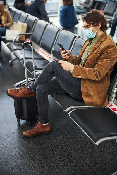 Stílusos férfi orvosi maszk segítségével mobiltelefon a repülőtéren — Stock Fotó