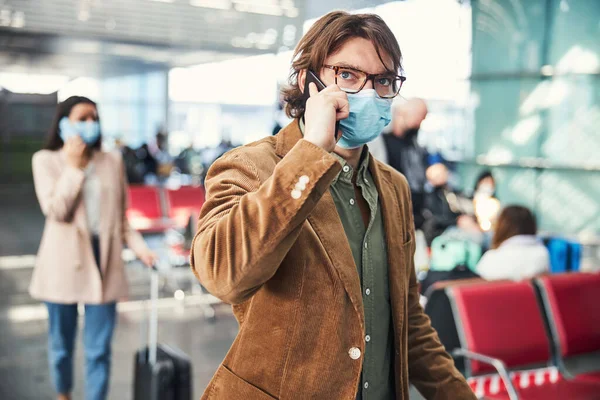 Egy fiatal férfi orvosi maszkban telefonál a reptéren. — Stock Fotó