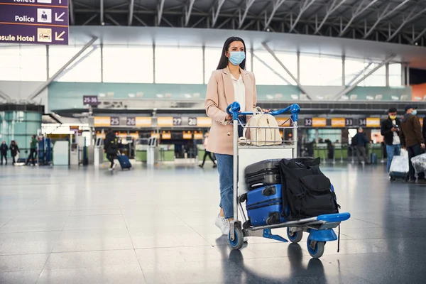 空港で荷物トロリーを押す医療マスクのスタイリッシュな女性 — ストック写真