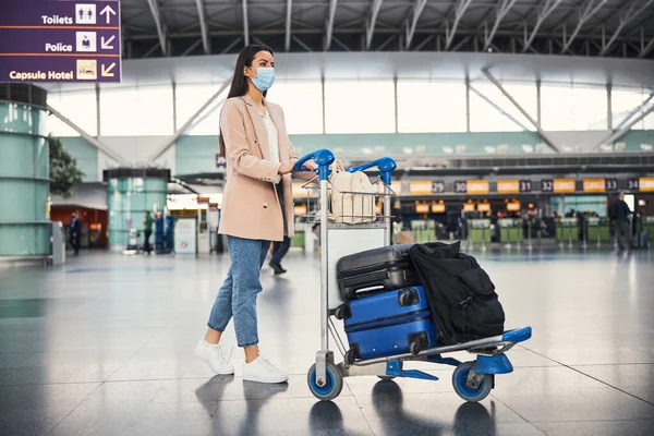空港で手荷物カートを押す医療マスクのスタイリッシュな女性 — ストック写真