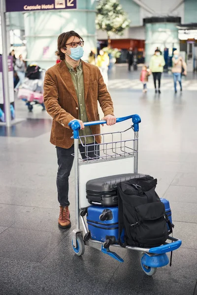 空港で手荷物カートを押す医療マスクのスタイリッシュな男 — ストック写真