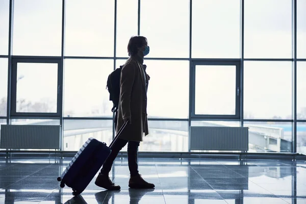 의료용 마스크를 입고 공항에서 여행 가방을 들고 있는 젊은 남자 — 스톡 사진