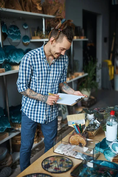 Vrolijke jongeman schrijft op klembord in werkplaats — Stockfoto