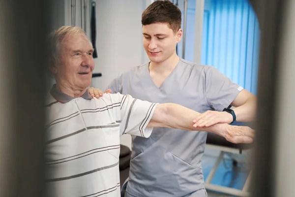 Fisioterapeuta que ayuda en el entrenamiento del brazo izquierdo del hombre anciano —  Fotos de Stock