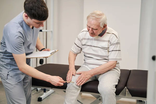 Fisioterapeuta apuntando su lápiz a la vieja rodilla del paciente — Foto de Stock