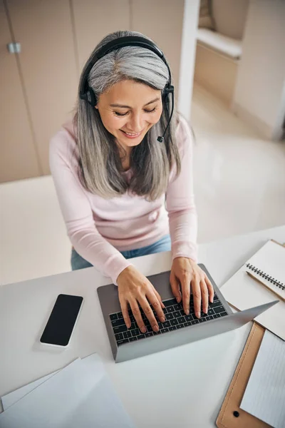 Charmig kvinna skrattar medan du sitter vid skrivbordet och arbetar på laptop — Stockfoto