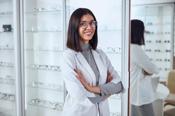Goed uitziende positieve vrouw in bril poseert in de kamer binnen in de voorkant van de stand met gereedschap voor het gezichtsvermogen — Stockfoto
