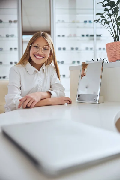 Cute girl in casual clothes sitting at the desk and looking at the camera in optician center — Stock Photo, Image