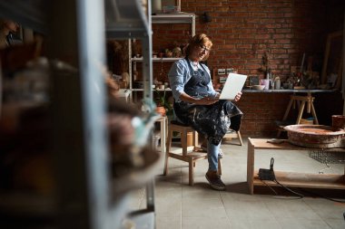 Çömlek atölyesinde dizüstü bilgisayar kullanan yaşlı hoş bir kadın.