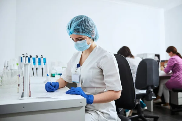 Bioquímica femenina con máscara facial escribiendo un informe de laboratorio de ciencias —  Fotos de Stock