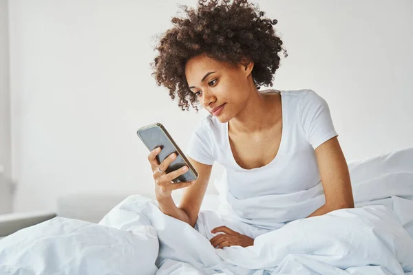 Γυναίκα με ένα smartphone στο χέρι κάθεται στο κρεβάτι — Φωτογραφία Αρχείου