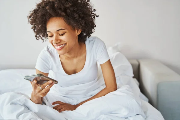 Ευτυχισμένο χαριτωμένο θηλυκό κάθεται στο κρεβάτι με το smartphone — Φωτογραφία Αρχείου