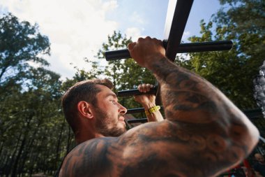 Kaslı dövmeli adam dışarıda barfiks çekiyor.