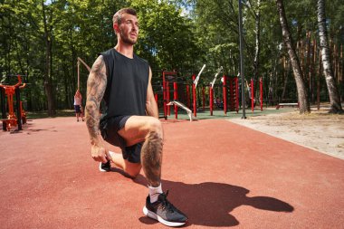 Motive olmuş sakallı adam spor sahasında bacak çalıştırıyor.