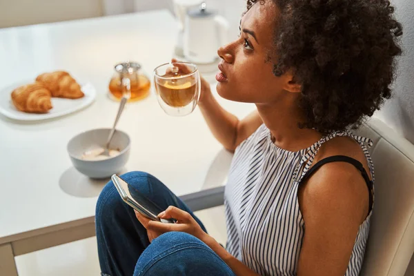 Жінка з напоєм і мобільний телефон за кухонним столом — стокове фото