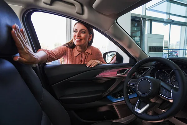 Joyeuse dame souriant en regardant à l'intérieur de la voiture et en touchant un volant — Photo