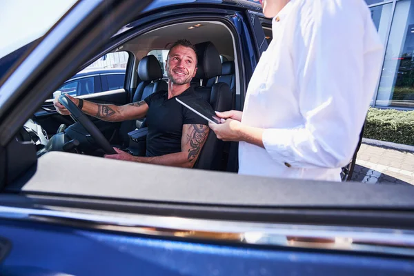 Gladsome tattooed man sitting in the car and smiling — Stock Photo, Image