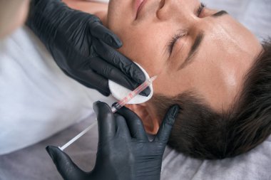 Kozmetik uzmanı koruyucu eldiven takıyor ve genç adama enjeksiyon yapıyor.