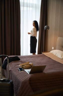 Otel misafiri pencerenin yanında duruyor, şık seyahat kıyafetleri giyiyor.