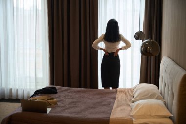 Otel misafiri bir otel odasında duruyor, yatağında dizüstü bilgisayar ve diğer şeyler var.