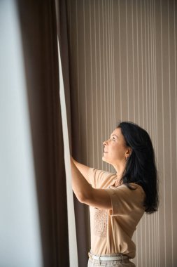 Kadın büyük bir pencere açıyor, rahat bir otel odasına yerleşiyor.