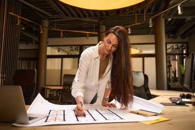 Konferans odasındaki bir masada çizimlerle çalışan bir kadın. Evin içinde, şık bir iç mekanda.