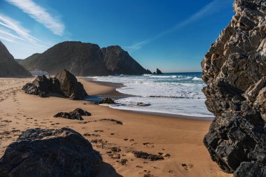 Ayak izleri Praia da Adraga 'daki koyu kıyı kayalıklarına ve mavi okyanus dalgalarına doğru gidiyor. Seyahat, manzara konsepti