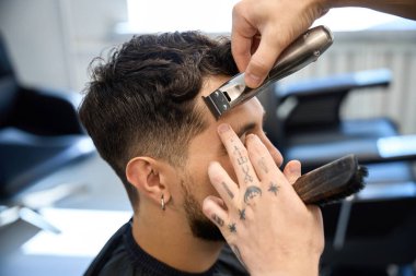 Profesyonel berberin yakın plan fotoğrafı, adamın yanında duruyor, elinde küçük bir saç makası ve saç kesme makinesi var.