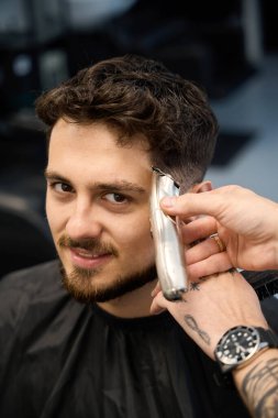 Profesyonel işçinin yakın plan fotoğrafı, yanında duran erkek, elinde küçük saç makası ve saç kesme makinesi var.
