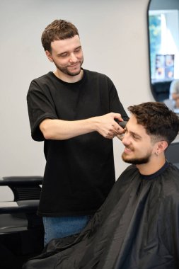 Profesyonel stilistin yakın plan fotoğrafı, yanında duran erkek, elinde saç makası ve saç kesme makinesi var.