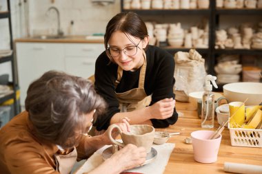 Yaşlı kadın bir atölyede kilden kase yapıyor. Bir ustanın gözetimi altında çalışıyor.