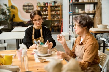 Genç bir kadın, işinde özel bir alet kullanır. Yaşlı bir kadın yanında oturur.