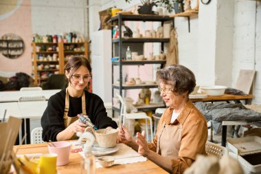 Kadın çömlekçi yanında yaşlı bir kadın otururken sprey tabanca kullanıyor.