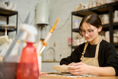 Kil örneğine özenle bakan genç bir kadın çömlek atölyesinde çalışıyor.