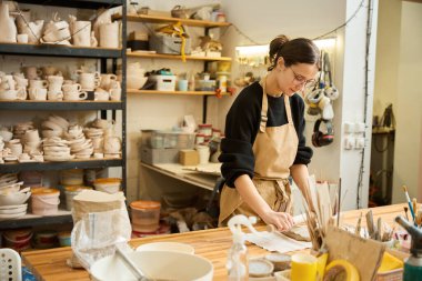 Genç bir kadın ahşap bir oklavayla kilden bir tabaka serer, her şey işyerinde düzenlenir.