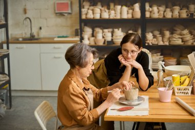Yaşlı bir kadın, özel aletler kullanarak bir zanaatkârın gözetiminde işlenmemiş kilden bir kase biçimlendiriyor.