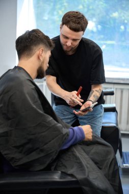 Yetenekli adam, modern berber dükkanında saç stili seçeneklerini telefonda gösteren müşterisiyle ayrıntılı istişarelerde bulunuyor.