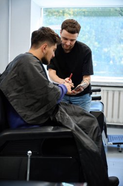 Yetenekli berber, modern berber dükkanında erkek saç kesimi seçeneklerini telefonda göstererek detaylı bir istişare içine girer.