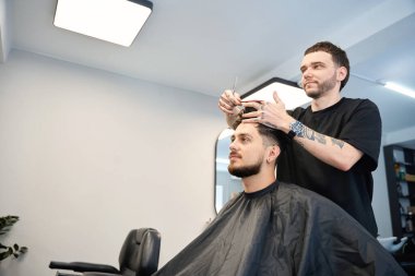 Profesyonel berber, elinde tarak ve makasla gülümseyen bir erkeğin yanında duruyor. Kesmeden önce saç uzunluğunu ölçüyor, aynaya bakıyor.