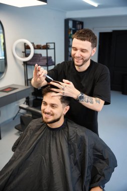 Profesyonel bir işçi, elinde tarak ve makasla gülümseyen bir erkeğin yanında duruyor, kesmeden önce saç uzunluğunu ölçüyor, aynaya bakıyor.