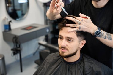 Profesyonel adam gülümseyen bir erkeğin yanında duruyor, elinde tarak ve makas, kesmeden önce saç uzunluğunu ölçüyor, aynaya bakıyor.