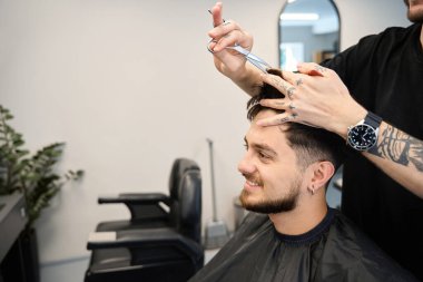 Profesyonel berber mutlu müşterinin yanında duruyor, elinde tarak ve makas, kesmeden önce saç uzunluğunu ölçüyor, aynaya bakıyor.