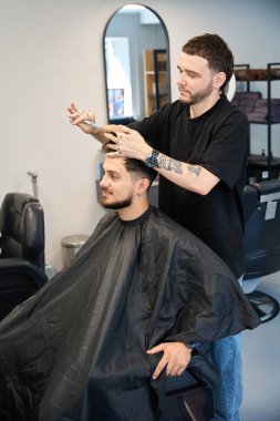 Profesyonel berber mutlu müşterinin yanında duruyor, elinde makasla, kesmeden önce saç uzunluğunu ölçüyor, aynaya bakıyor.