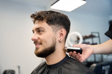 Profesyonel berber gülümseyen adamın yanında duruyor, elinde traş makinesi, saçlarını kesiyor, saçını kesiyor.