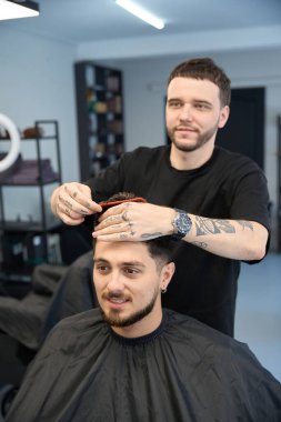 Profesyonel berber gülümseyen adamın yanında duruyor, elinde tarak tutuyor ve kesmeden önce saç uzunluğunu ölçüyor.