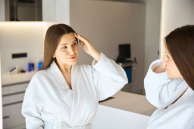 Beyaz bornozlu gülümseyen genç bayan güzellik salonunda aynaya bakıyor. Sağlık Merkezi 'nde. Sağlık, klinik, kozmetoloji