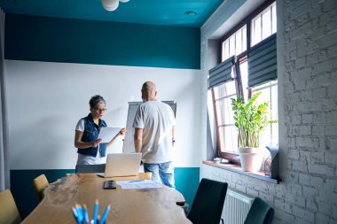 Gri saçlı ve gözlüklü olgun bir kadın ofiste kel bir adamla tartışıyor. Doğal ışık ve işbirlikçi ruhla güçlendirilmiş odaklanmış atmosfer.